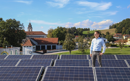 Mann steht stolz auf seinem Dach zwischen vielen Photovoltaikmodulen. Im Hintergrund sieht man Teile des Dorfes Baumgartenberg in Oberösterreich.