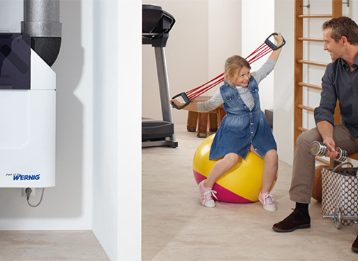 Wohnraumlüftungsgerät in offenem Abstellraum, im Fitnessraum daneben spielt ein Mann mit einem Kind.