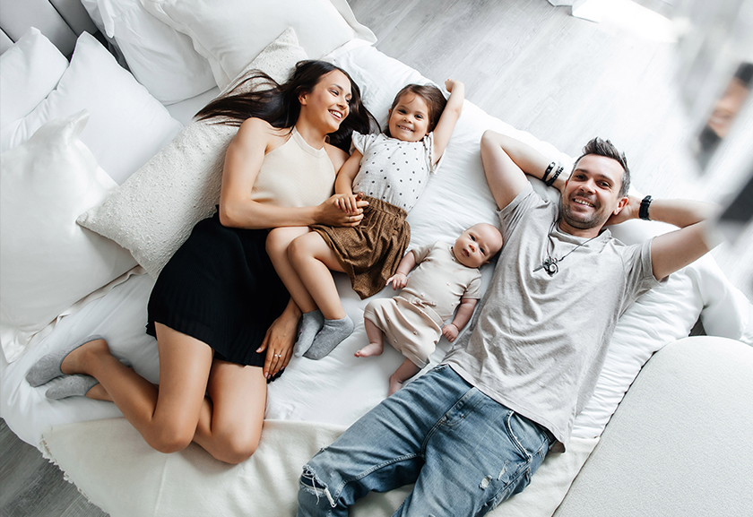 Eine junge Frau, zwei Kinder und ein Mann liegen auf einem hell überzogenen Bett und genießen die gute Luft.